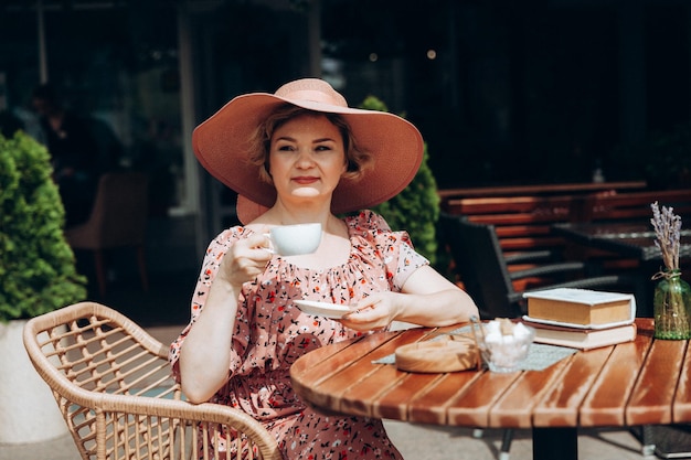 Retrato de moda ao ar livre de uma mulher deslumbrante sentada em um café eu bebo café e leio um livro antigo uma mulher de vestido e chapéu