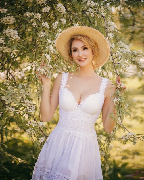 Retrato de moda arte de uma bela jovem loira em um jardim de verão verde florescendo em um vestido de luz branca, com um chapéu de palha. Garota no estilo country