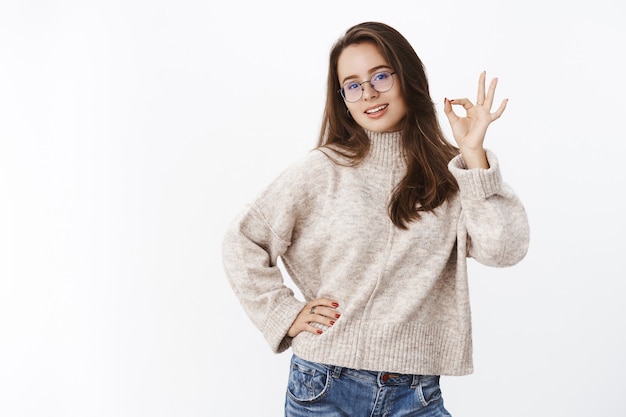 Retrato de modelo feminino elegante e autoconfiante, com cabelo castanho de óculos e suéter segurando a mão no quadril, mostrando um gesto de ok ou ok em confirmação, gosto ou aprovação dando feedback positivo.