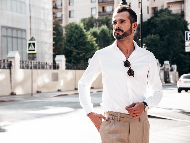 Retrato de modelo lambersexual bonito confiante elegante hipster homem moderno sexy vestido com camisa branca e calças Moda masculina posando no fundo da rua na cidade da Europa ao pôr do sol em óculos de sol