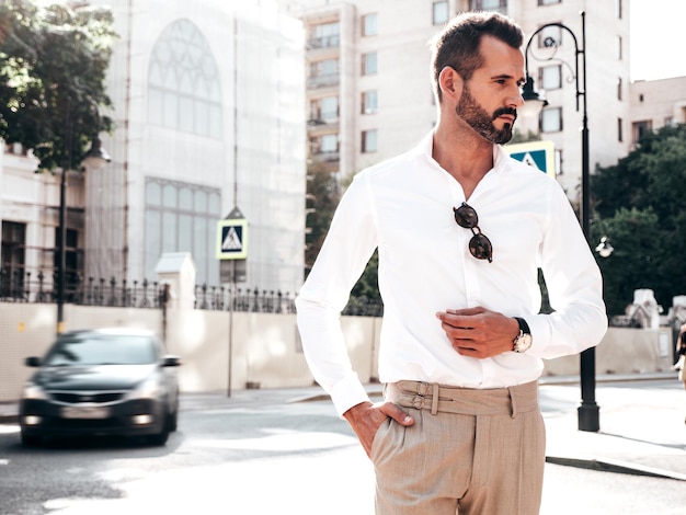 Retrato de modelo lambersexual bonito confiante elegante hipster homem moderno sexy vestido com camisa branca e calças Moda masculina posando no fundo da rua na cidade da Europa ao pôr do sol em óculos de sol