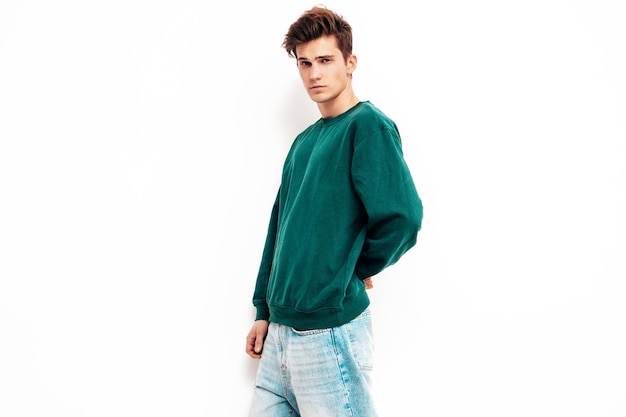 Retrato de modelo lambersexual bonito e elegante hipster homem vestido de jeans e suéter verde Moda masculina posando no estúdio perto da parede branca