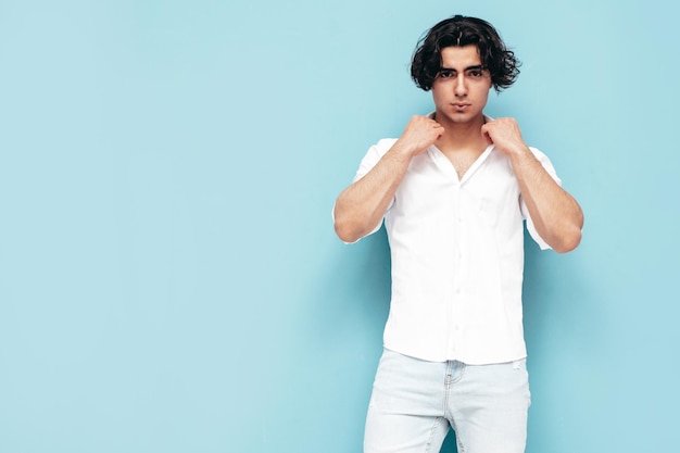 Retrato de modelo lambersexual hipster elegante e confiante bonito Homem sexy vestido com camisa branca de verão e roupas jeans Moda masculina isolada no estúdio posando perto da parede azul