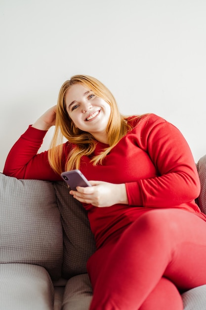 Retrato de modelo sorridente de tamanho maior sentado no sofá na sala de estar usando telefone celular olhando para a câmera mensagem de texto Conceito de tecnologia