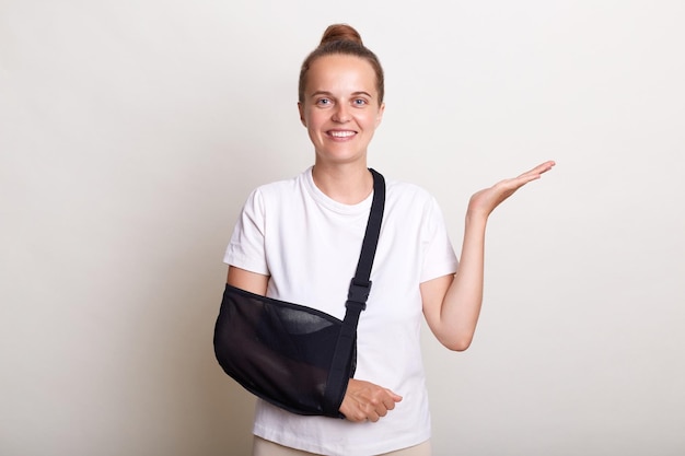 Foto retrato de mulher adorável satisfeita com penteado de coque mantém a mão espalhada tem expressão sorridente vestida de branco camiseta se recupera após acidente ter quebrado o braço usa estilingue após visitar cirurgião