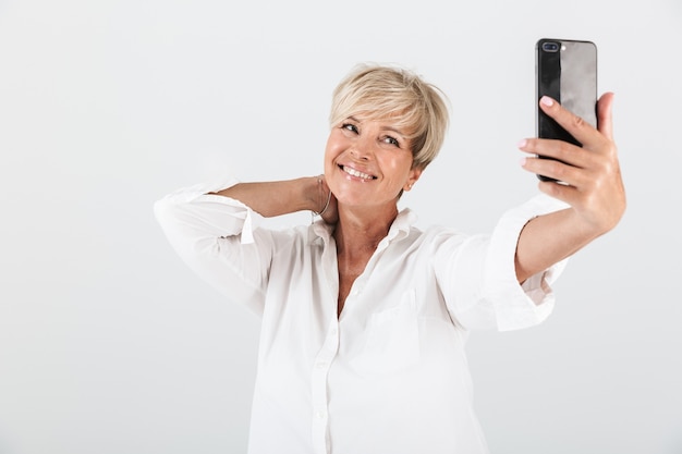 Foto retrato de mulher adulta alegre com cabelo loiro curto, sorrindo e tirando selfie retrato no celular isolado sobre uma parede branca em estúdio