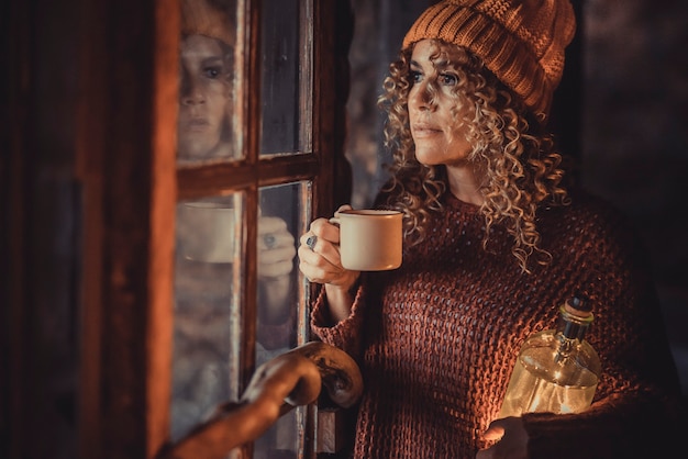 Retrato de mulher adulta atraente em pé em casa e olhando pela janela com pensamentos. atmosfera e luz do inverno do feriado. pessoas femininas de expressão bem séria