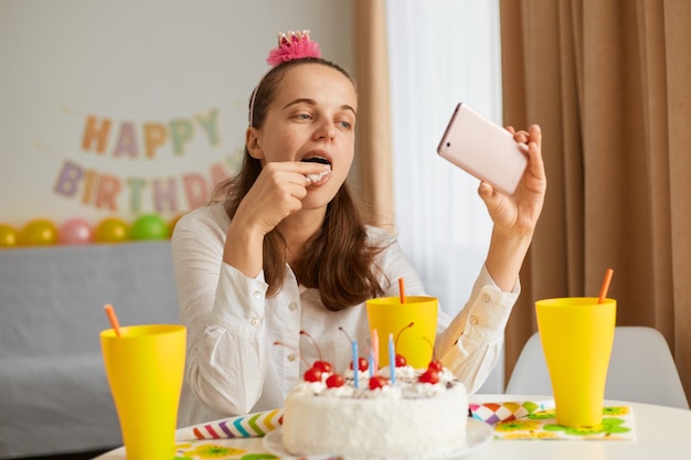 Retrato de mulher adulta jovem faminta vestindo traje branco, sentado à mesa com bolo e bebida, segurando o celular nas mãos, saboreando uma deliciosa sobremesa e tomando via videochamada.