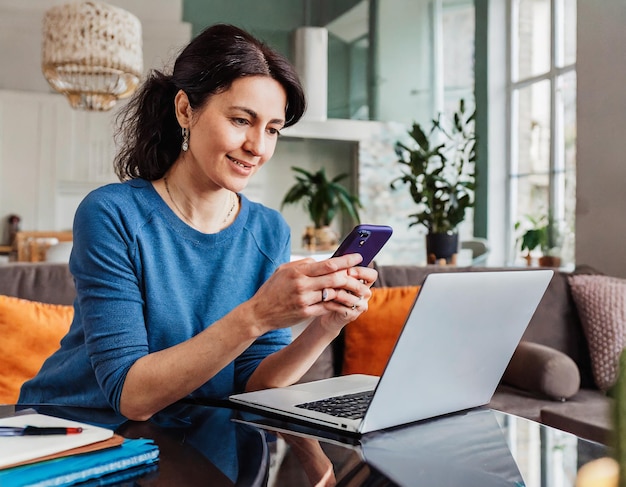 Retrato de mulher adulta usando telefone móvel enquanto está sentada na sala de estar com um laptop