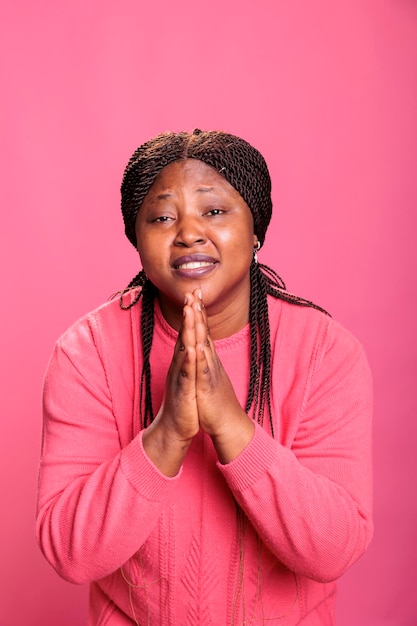 Retrato de mulher afro-americana religiosa orando a deus pedindo perdão em pé no estúdio com fundo rosa. jovem adulto segurando as palmas das mãos juntas em oração. conceito de religião