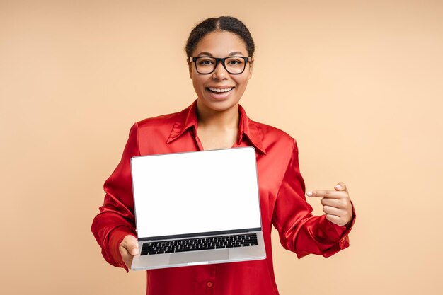 Foto retrato de mulher afro-americana sorridente segurando um laptop apontando o dedo para a tela branca olhando para a câmera isolada em fundo bege