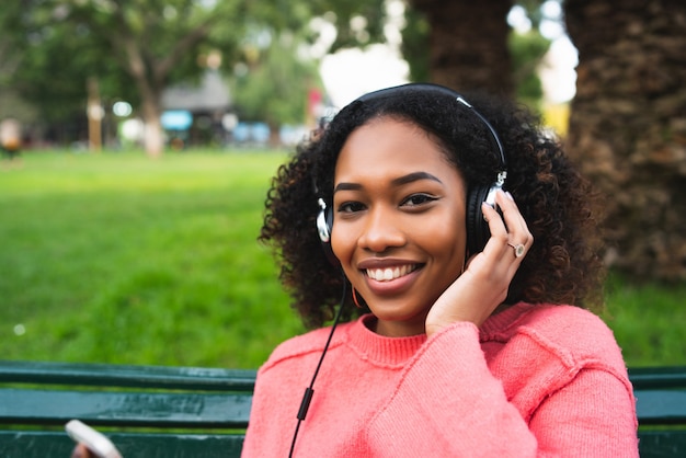 Retrato de mulher afro-americana, sorrindo e ouvindo música com fones de ouvido no parque. Ao ar livre.