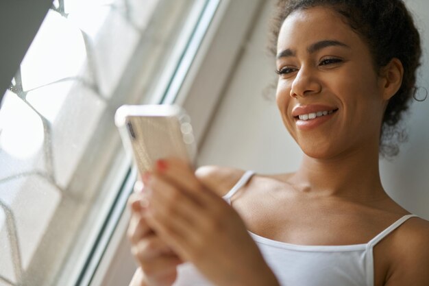 Retrato de mulher alegre jovem mestiça sorrindo, usando smartphone, sentindo-se confortável enquanto está sentado perto da janela em casa