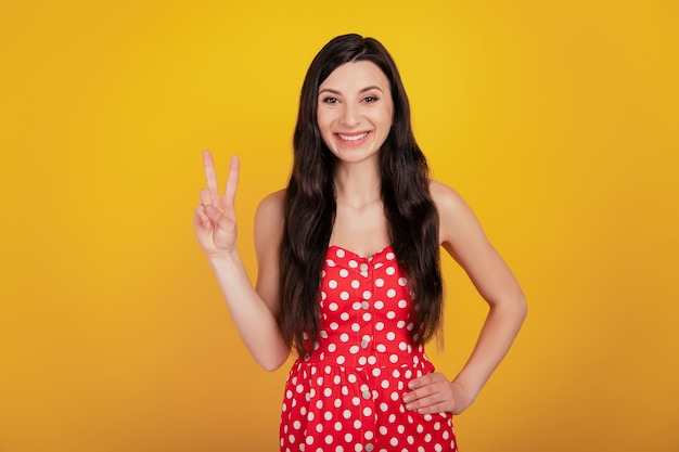 Retrato de mulher alegre mostra sinal de V sorriso branco e usa um vestido vermelho pontilhado sobre fundo amarelo