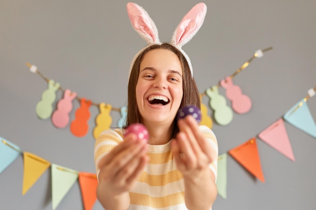 Retrato de mulher alegre rindo usando orelhas de coelho segurando dois ovos de páscoa coloridos mostrando seus ovos pintados à mão estando de bom humor festivo isolado em fundo cinza decorado