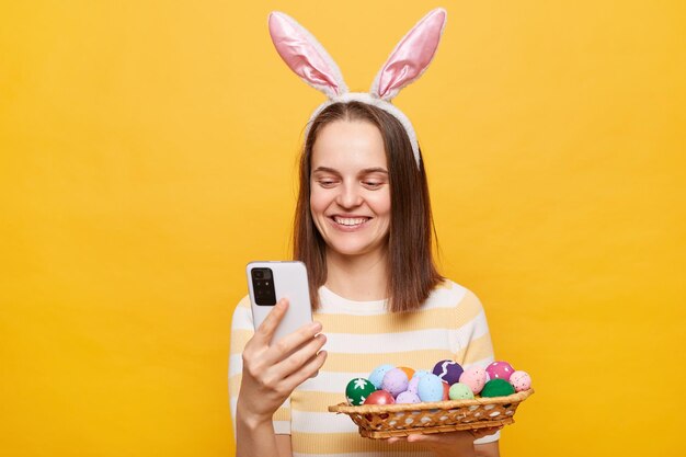 Retrato de mulher alegre sorridente usando orelhas de coelho segurando ovos de páscoa isolados sobre fundo amarelo usando celular massageando com amigos parabenizando com feriado