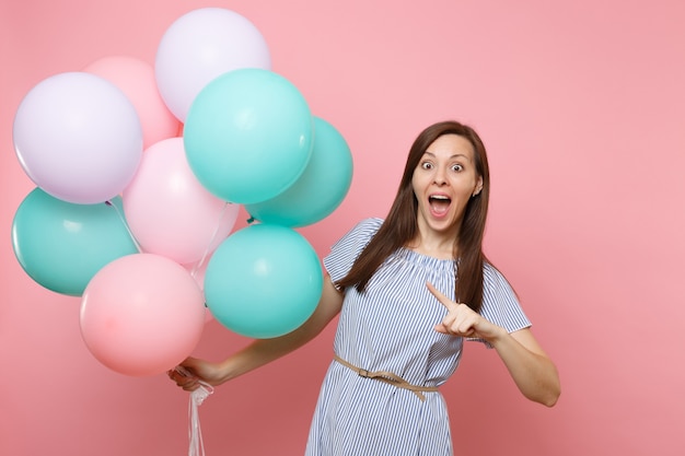 Retrato de mulher alegre surpresa com a boca aberta no vestido azul, apontando o dedo indicador em balões de ar coloridos isolados no fundo rosa. festa de aniversário, conceito de emoções sinceras de pessoas.