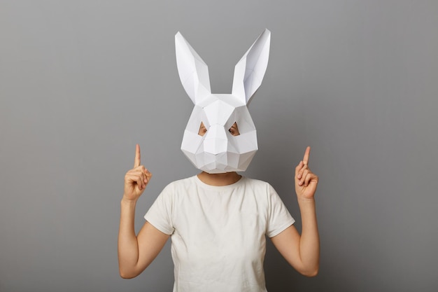 Retrato de mulher anônima vestindo camiseta branca e máscara de coelho de papel em pé isolado sobre fundo cinza, apontando os dois dedos indicadores para cima e copie o espaço para o texto promocional