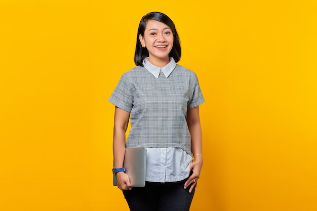 Retrato de mulher asiática alegre trazendo laptop sobre fundo amarelo