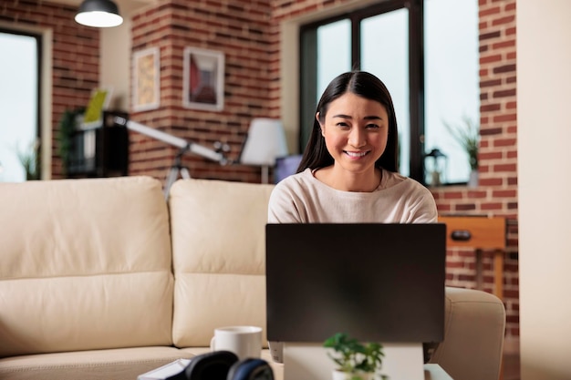 Retrato de mulher asiática bonita sorridente olhando direto para a câmera na moderna sala de estar plana. tecnologia laptop internet estilo de vida feliz trabalho em casa freelancer