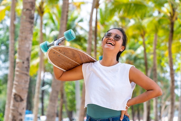 Retrato de mulher asiática sorridente atraente segurando um skate caminhando na praia em um dia ensolarado de verão Confiança feminina desfrutar e se divertir com atividades ao ar livre estilo de vida e esportes extremos skate de surf