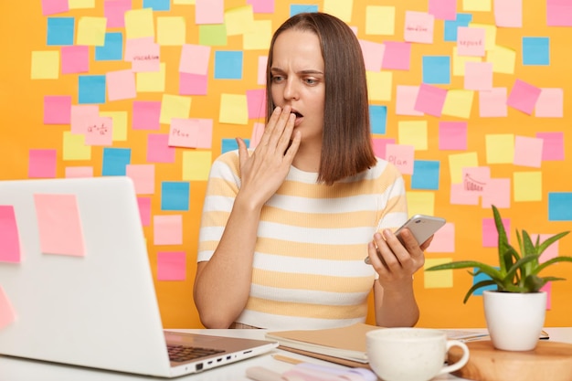 Retrato de mulher assustada chocada com cabelo castanho vestindo camiseta listrada sentada à mesa olhando para o monitor do laptop cobrindo a boca tem erro posando contra a parede amarela com pequenas notas adesivas