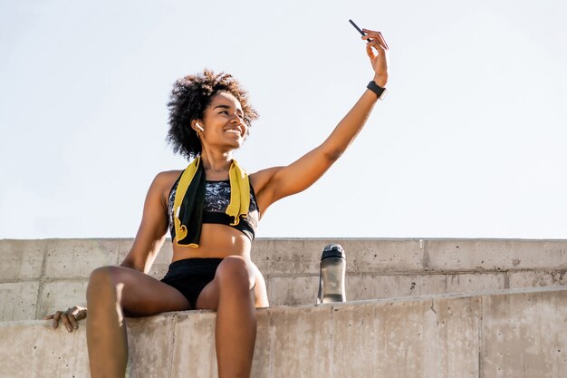 Foto retrato de mulher atleta afro tirando uma selfie com o celular e relaxando depois do treino ao ar livre