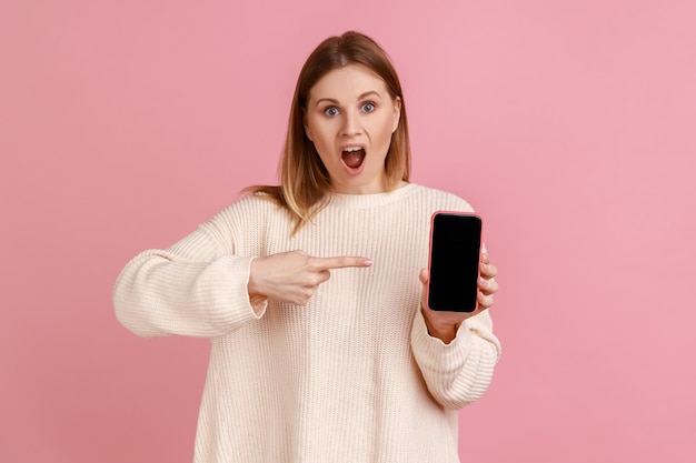 Retrato de mulher atônita segurando o celular nas mãos, apontando para a tela em branco do smartphone com espaço de cópia para promoção, vestindo suéter branco. Tiro de estúdio interior isolado no fundo rosa.