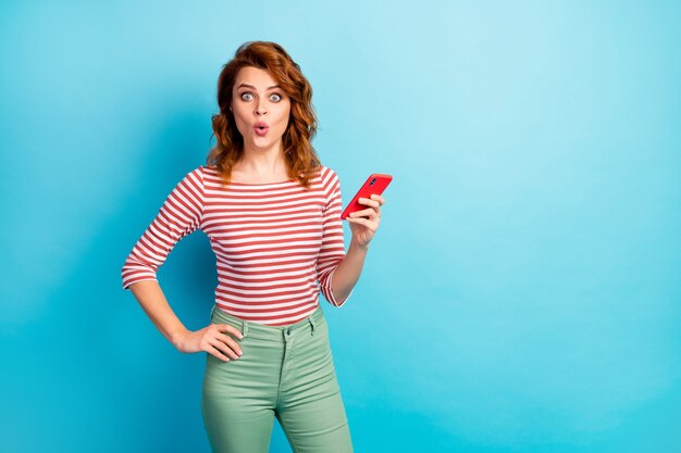 Retrato de mulher atônita, usar telefone inteligente, receber notificação de mídia social, gritar impressionado, uau, usar um suéter de boa aparência isolado sobre a cor azul