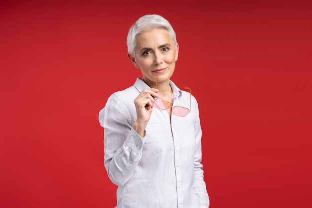 Foto retrato de mulher atraente de cabelos grisalhos segurando óculos de sol olhando para a câmera isolada em fundo vermelho bela mulher de negócios em camisa rosa elegante funcionária bem sucedida conceito de negócio