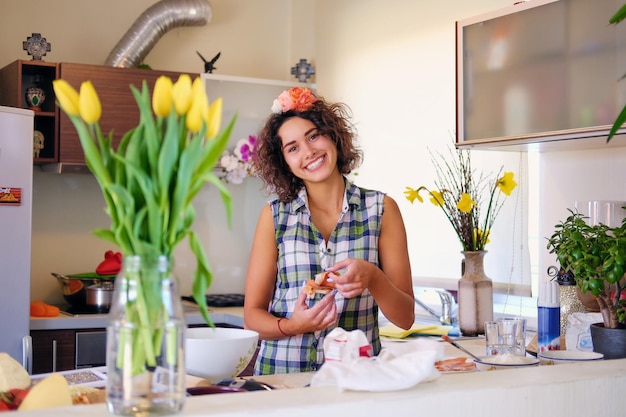 Retrato de mulher atraente morena com cabelo encaracolado em uma cozinha de casa com muitas flores.