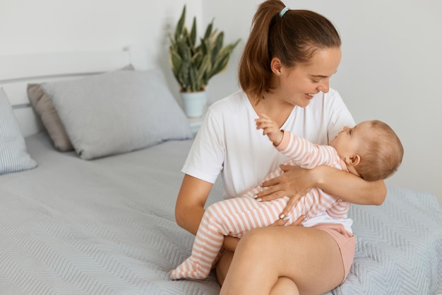 Retrato de mulher atraente satisfeita feliz vestindo camiseta branca e shorts sentado na cama com sua filha bebê, mãe brincando com seu filho, expressando amor e gentil.