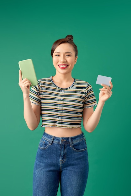 Retrato de mulher atraente segurando telefone celular e cartão de crédito nas mãos isoladas sobre fundo verde