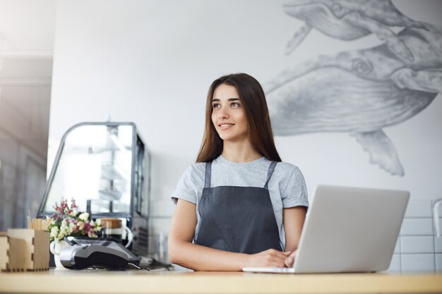 Retrato de mulher barista tendo um breve descanso de seu trabalho no café ocupado, aproveitando a luz do sol usando um laptop.