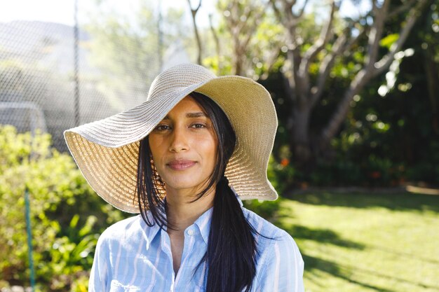 Retrato de mulher biracial feliz com chapéu de sol em pé no jardim ensolarado sorrindo. Conceito de felicidade, saúde, vida doméstica e inclusão.