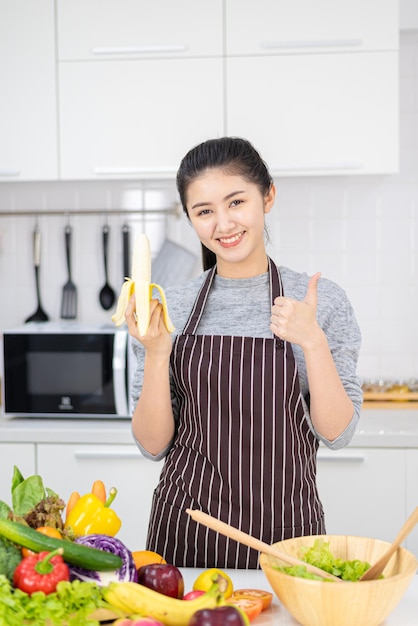 Retrato de mulher bonita asiática e dona de casa está sorrindo e segurando banana na cozinha branca