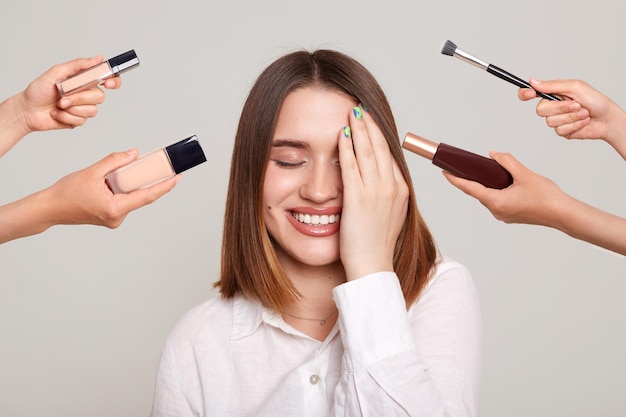 Retrato de mulher bonita atraente com maquiagem em pé com sorriso e olhos fechados cobrindo o olho com a palma cercada com as mãos de várias esteticistas segurando seus respectivos equipamentos