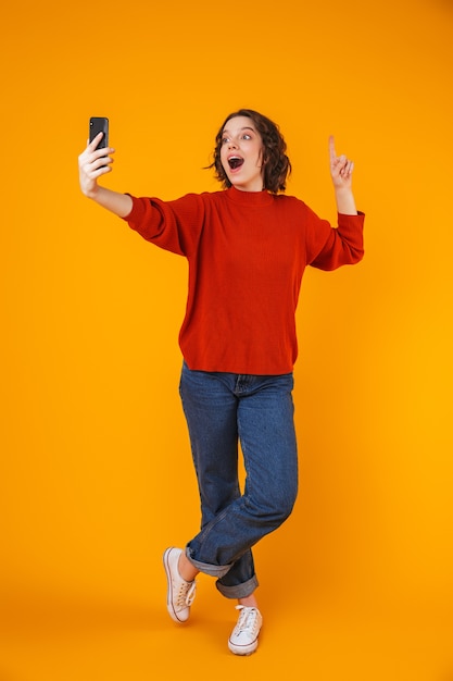 retrato de mulher bonita jovem emocional animado posando isolado sobre uma parede amarela tome selfie pelo telefone móvel.