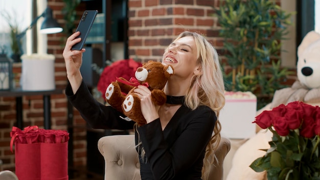 Retrato de mulher bonita no dia dos namorados segurando smartphone tomando selfie com ursinho de presente de um ente querido. loira sedutora sorridente em videochamada sentindo-se amada na sala com presentes de luxo.