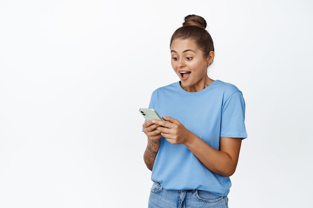 Retrato de mulher bonita parece surpreso com a tela do celular, garota usando smartphone e sorrindo espantada com branco