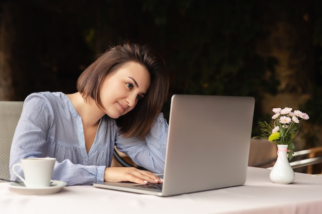 Retrato de mulher bonita que trabalha em casa, ela se senta com uma xícara de café na mesa, trabalhando no laptop dentro de casa