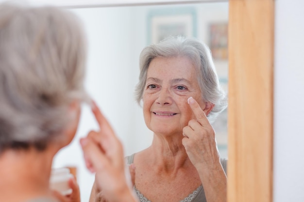 Retrato de mulher bonita sênior aplica creme antienvelhecimento no rosto enrugado senhora sorridente idosa segurando um produto de frasco cosmético