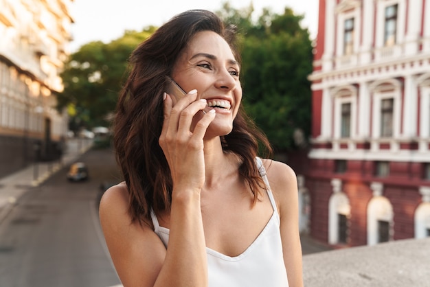 Retrato de mulher bonita sorrindo e falando no smartphone em pé na ponte com a rua da cidade