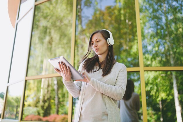 Foto retrato de mulher bonita, vestindo roupas leves casuais. menina segurando o computador tablet pc, ouvindo música em fones de ouvido no parque da cidade, perto do espelho, construindo ao ar livre na natureza da primavera. conceito de estilo de vida.