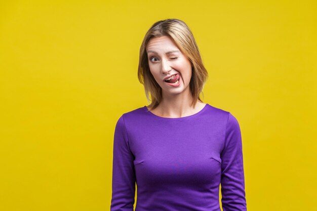 Retrato de mulher brincalhão alegre em vestido roxo apertado, piscando e enfiando a língua para fora, mostrando uma careta de rosto engraçado, se divertindo na câmera, tiro de estúdio interno, isolado em fundo amarelo