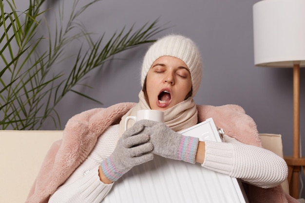 Retrato de mulher cansada com sono de casaco e chapéu sentado na casa fria sente congelar tentando se aquecer com aquecimento elétrico e chá quente bocejando com os olhos fechados