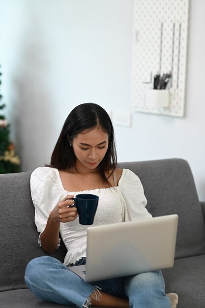 Retrato de mulher casual usando laptop e bebendo café enquanto está sentado no sofá em casa.