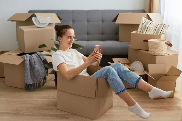 Retrato de mulher caucasiana de cabelos escuros feliz vestindo camiseta branca e jeans sentado em uma caixa de papelão segurando o celular e rolando on-line realocando em seu novo apartamento