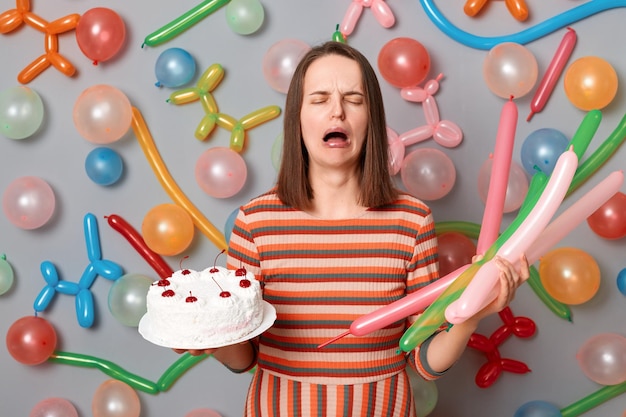 Retrato de mulher caucasiana desesperada com cabelo castanho usando vestido listrado em pé ao redor de balões inflados multicoloridos segurando bolo e chorando sente-se sozinha em seu aniversário