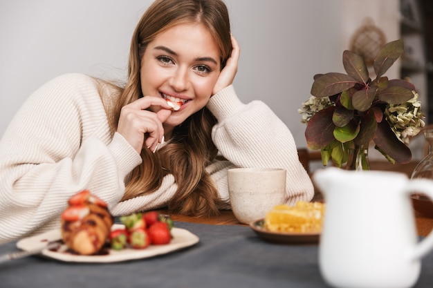 retrato de mulher caucasiana feliz vestindo roupas casuais, comendo morango enquanto toma o café da manhã em um quarto aconchegante